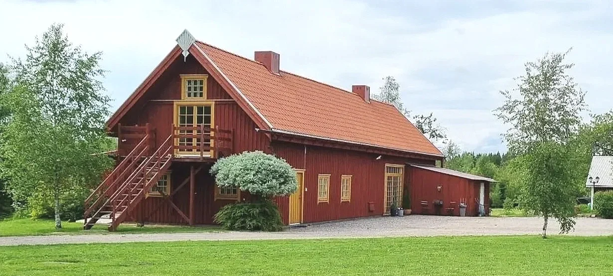 Ein rotes Holzhaus mit Steinterrasse auf einer grünen Wiese, umgeben von Bäumen und einem Weg davor.