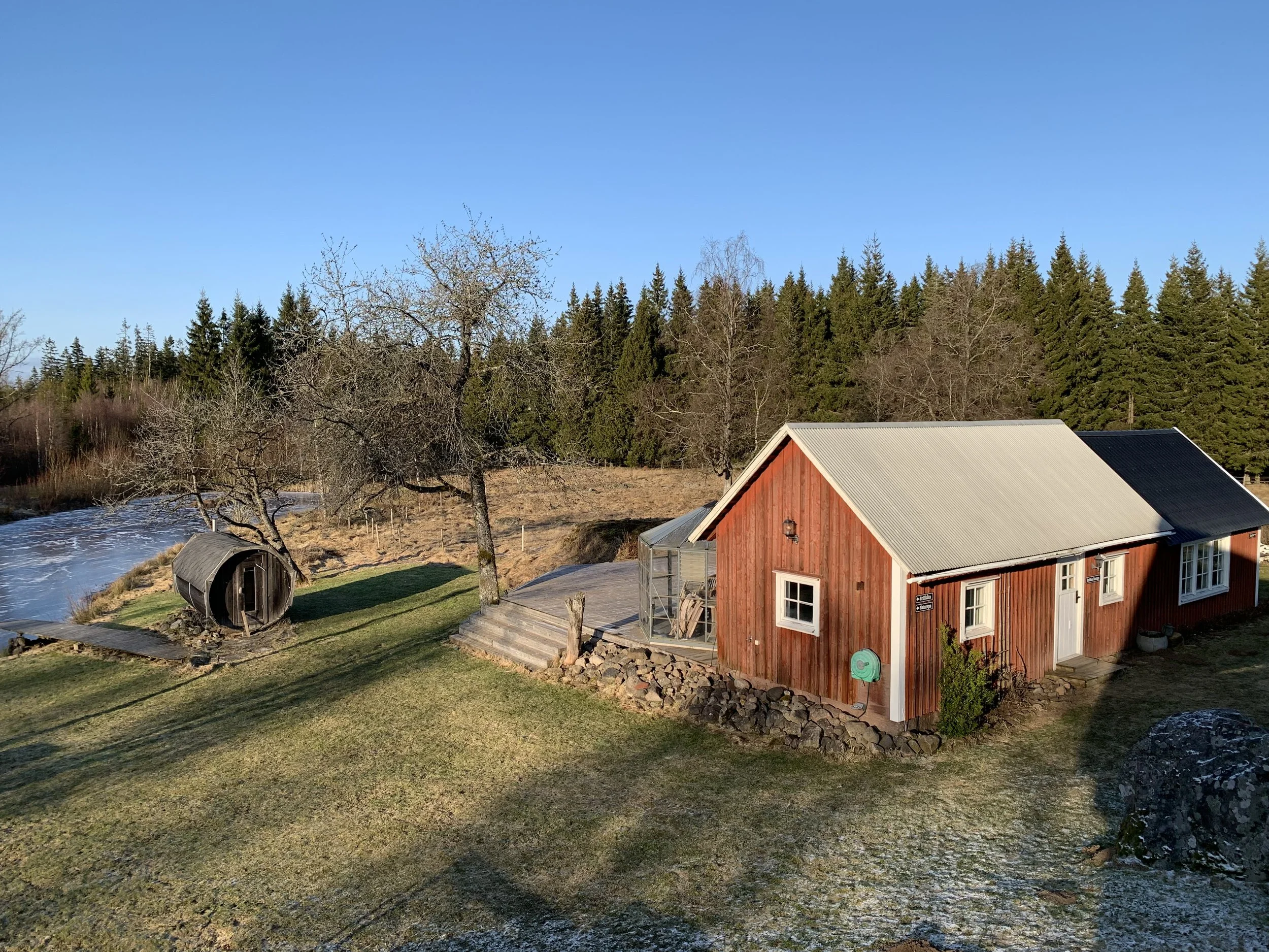 Ein rotes Holzhaus mit weißen Fenstern inmitten einer ländlichen Landschaft, umgeben von Bäumen und Wiesen, bei sonnigem Wetter.
