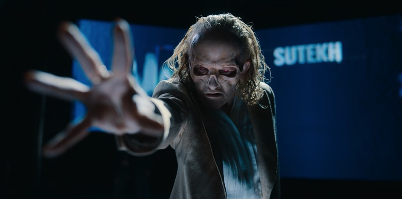 Still from Doctor Who. A person with zombie-like makeup and long hair reaches out towards the camera in a dark room. There is a blue screen in the background with the text "SUTEKH" visible.