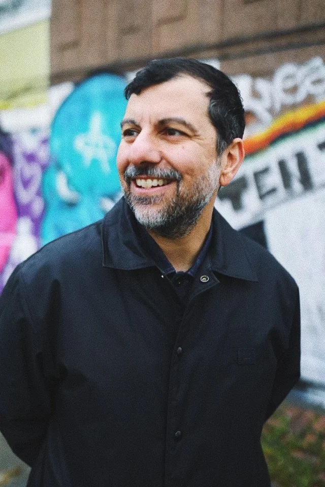 Huse Mondaradi, wearing a black jacket, standing in front of colorful graffiti on a brick wall.
