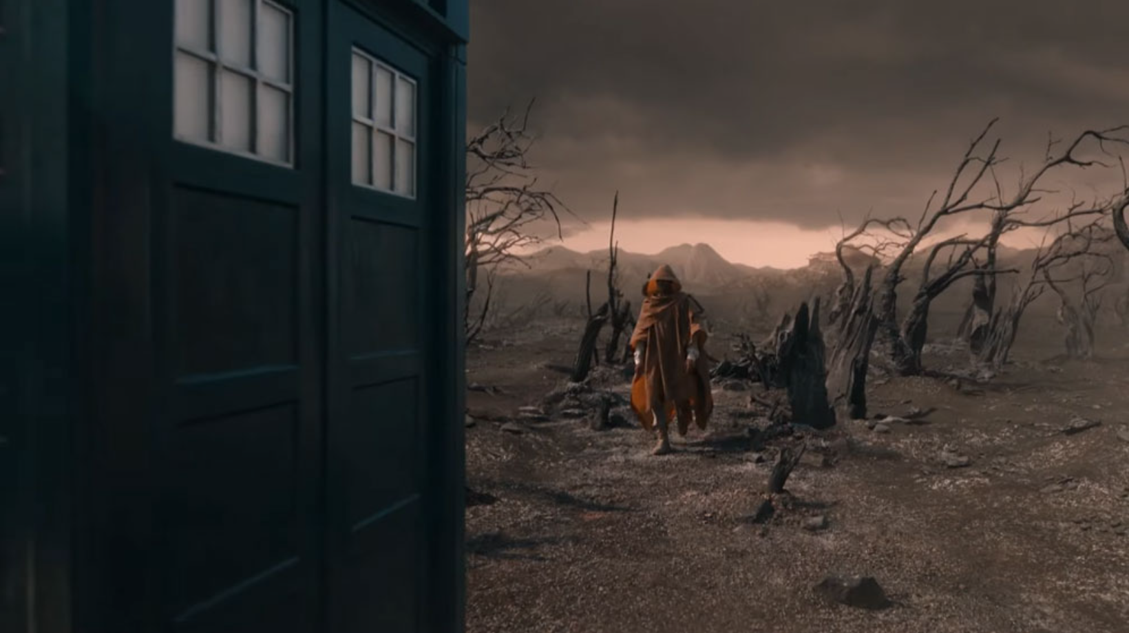 Still from Doctor Who. A post-apocalyptic, barren landscape with dead trees and mountains in the background, under a dark, cloudy sky.