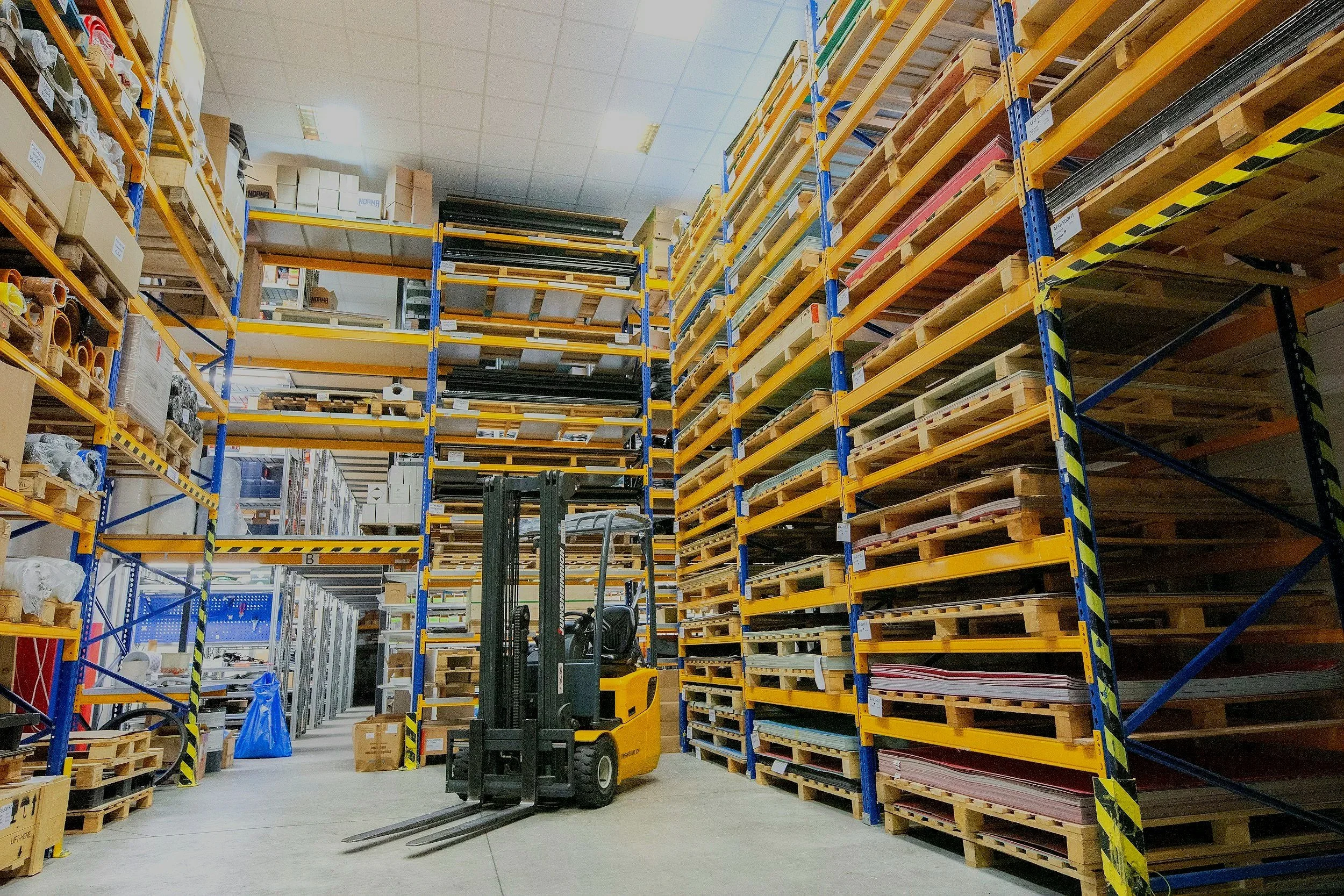 Interior of a warehouse with tall shelving units filled with boxes and materials, a forklift parked in the aisle.
