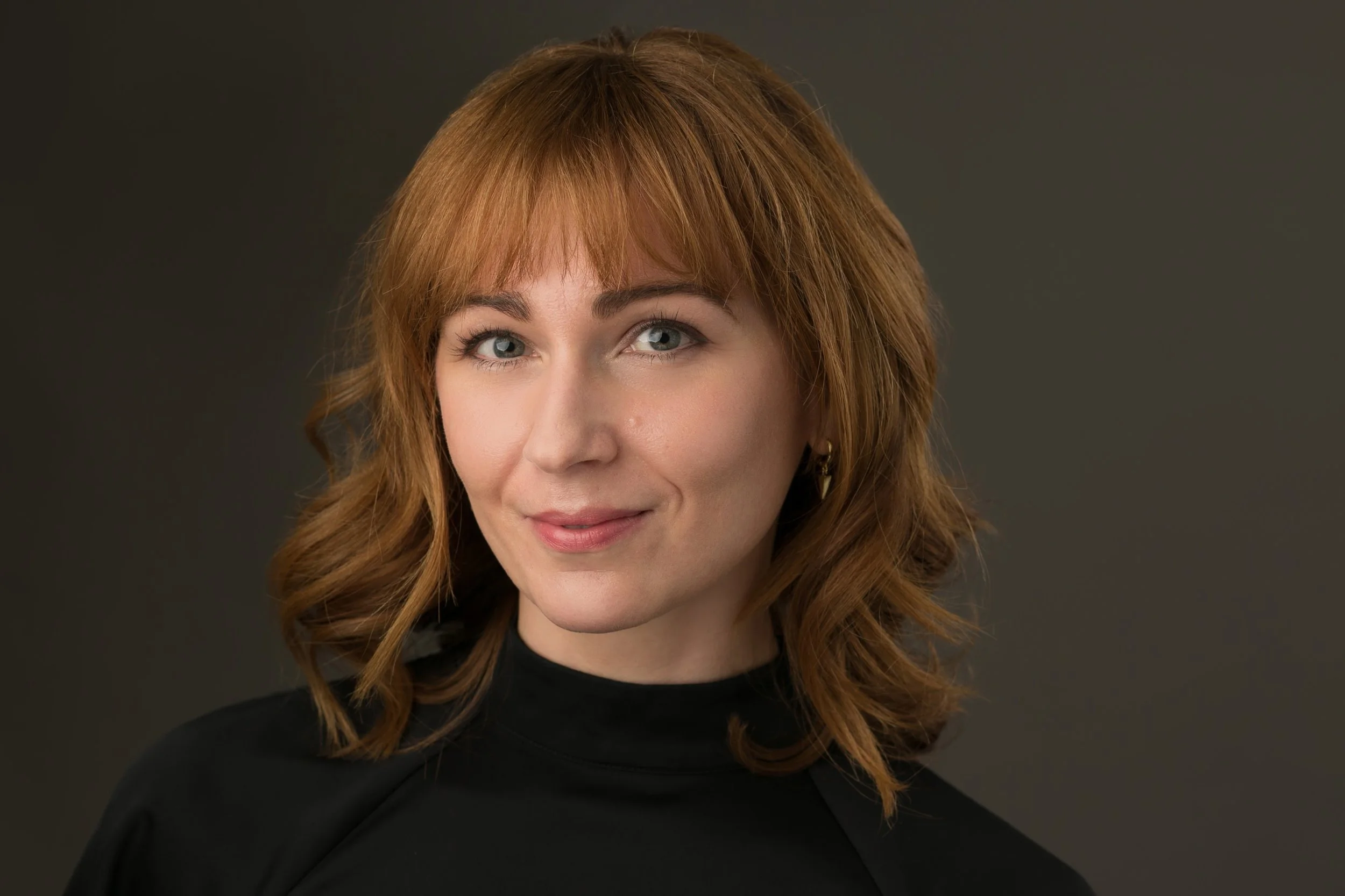 A woman with shoulder-length red hair and blue eyes, wearing a black top, smiling slightly at the camera with a dark gray background.
