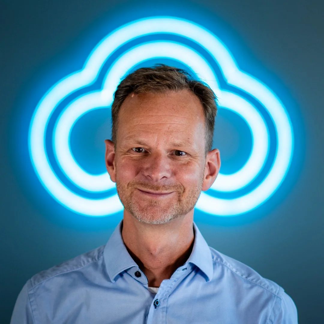 A man with short brown hair and a light beard, wearing a light blue shirt, standing in front of a blue cloud-shaped neon sign.