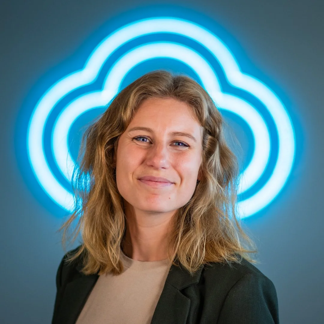 Portrait of a smiling woman with shoulder-length wavy blonde hair, wearing a beige top and a black jacket, standing in front of a blue neon cloud-shaped sign.
