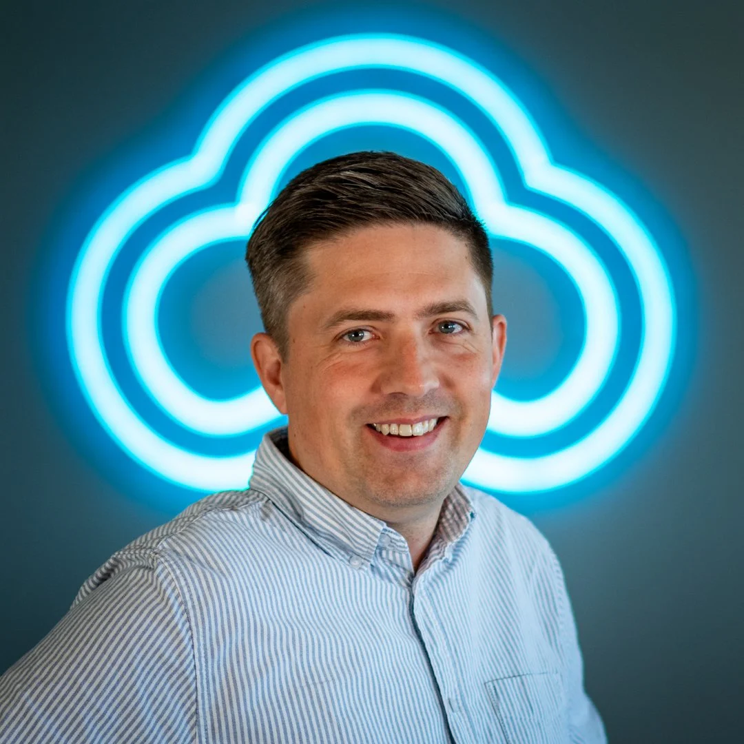 A man with short dark hair smiling, wearing a striped button-up shirt, standing in front of a illuminated cloud-shaped neon sign.