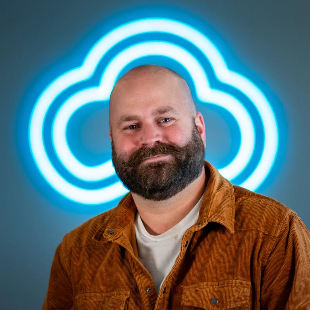 A man with a beard and a shaved head smiling, wearing a brown shirt over a white t-shirt, standing in front of a blue neon cloud-shaped sign.