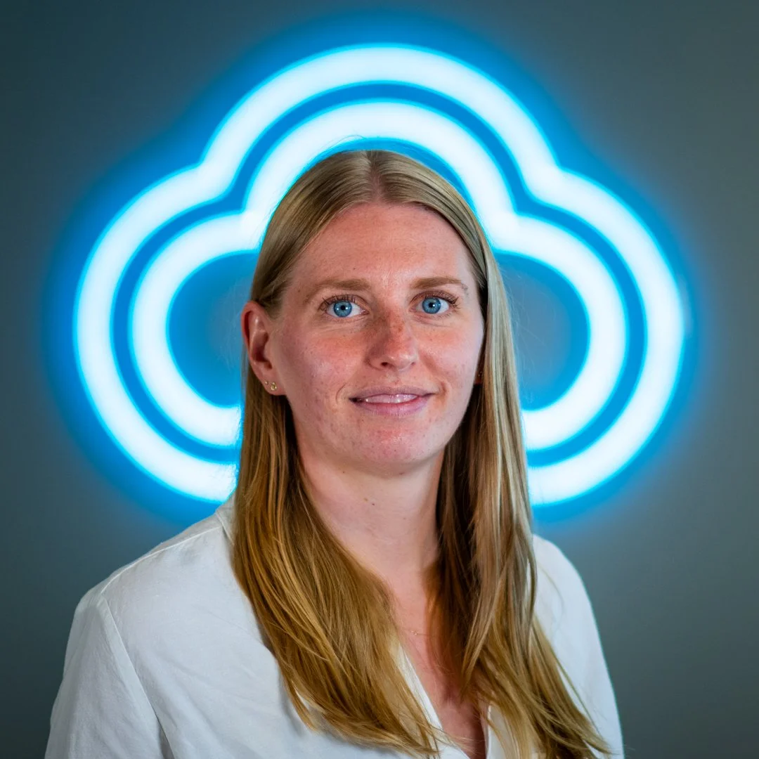 Portrait of a woman with blonde hair and blue eyes in front of a glowing blue cloud-shaped neon sign.