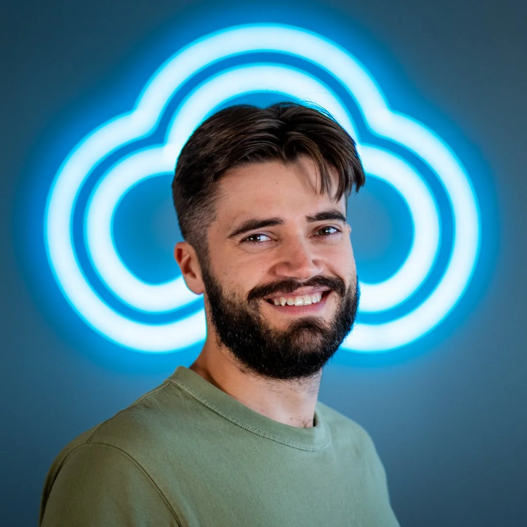A smiling man with a beard and short brown hair, wearing a green shirt, standing in front of a neon blue cloud-shaped logo on a dark wall.
