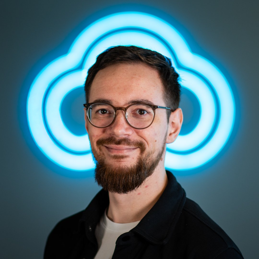 A man with glasses and a beard smiling in front of a glowing blue cloud logo.