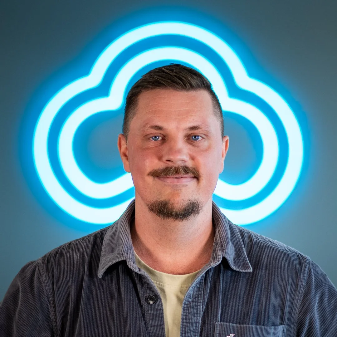 A man with a mustache and goatee, wearing a gray shirt, standing in front of a blue neon cloud logo.