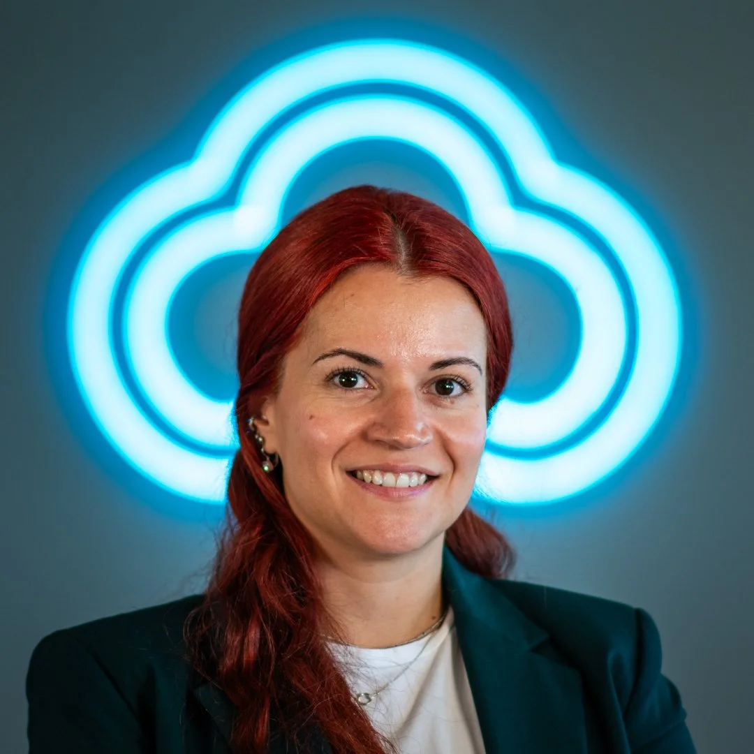A woman with red hair smiling in front of a blue neon cloud-shaped sign.