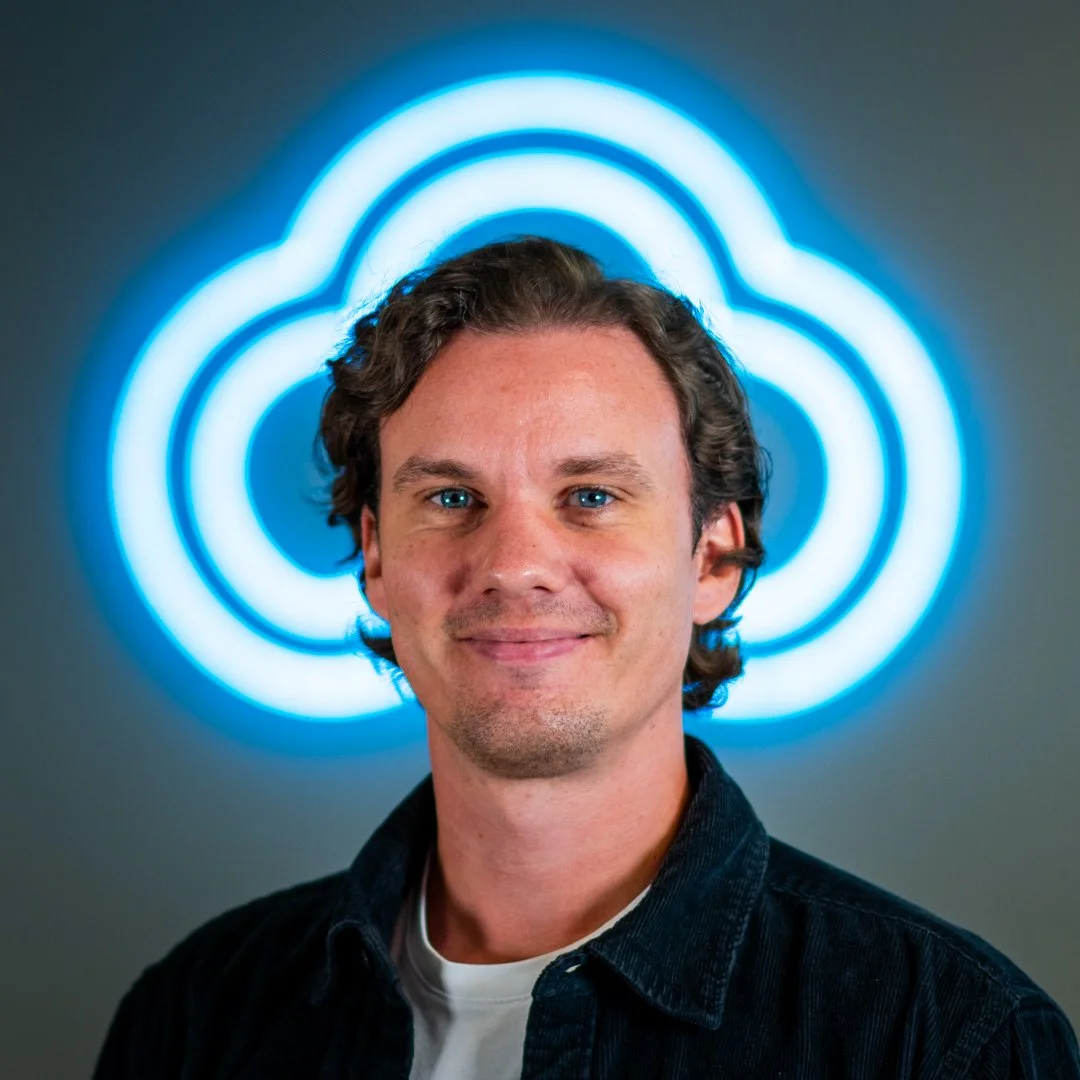 A man with curly brown hair and blue eyes smiling in front of a neon cloud logo.