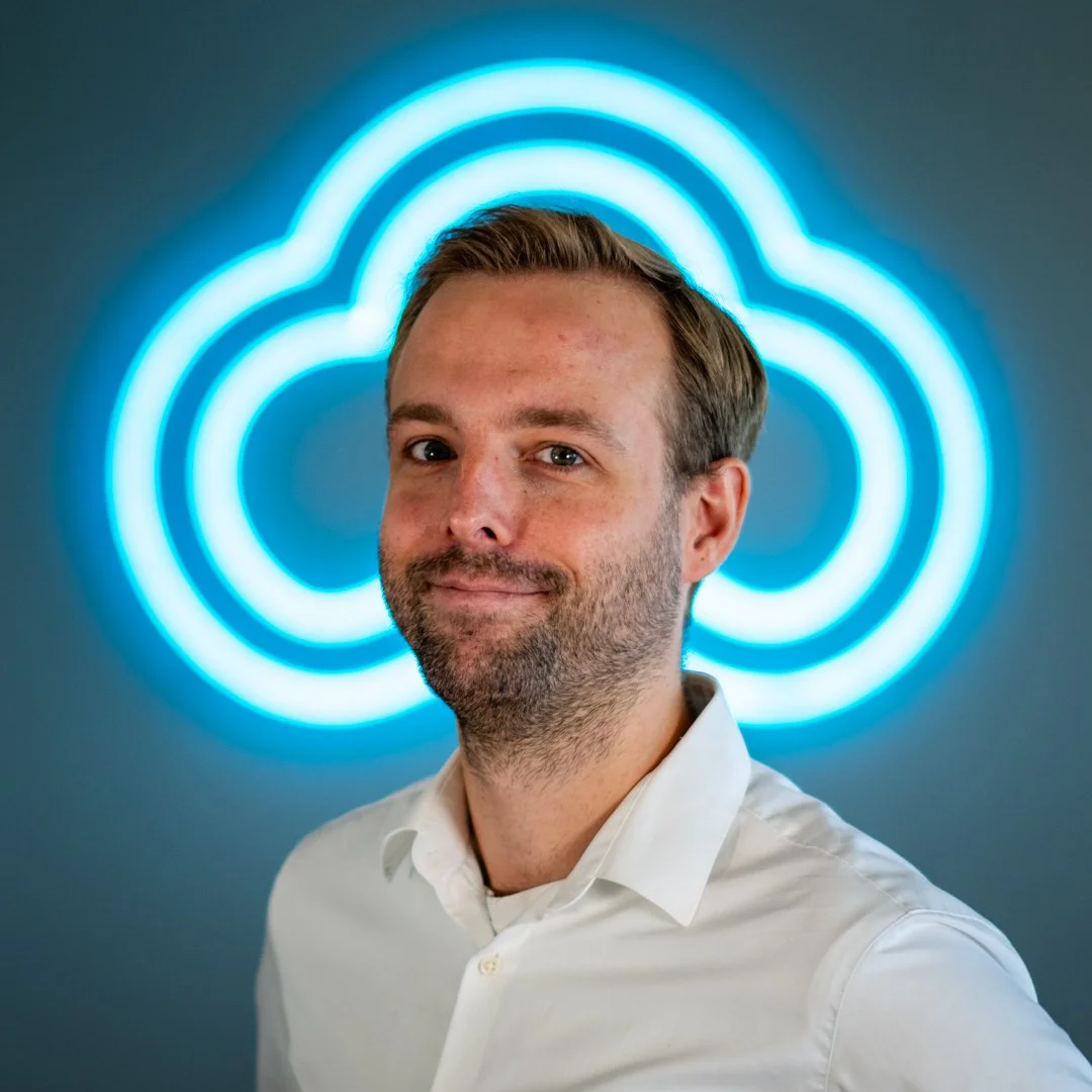 A man in a white shirt standing in front of a neon cloud logo on a wall.