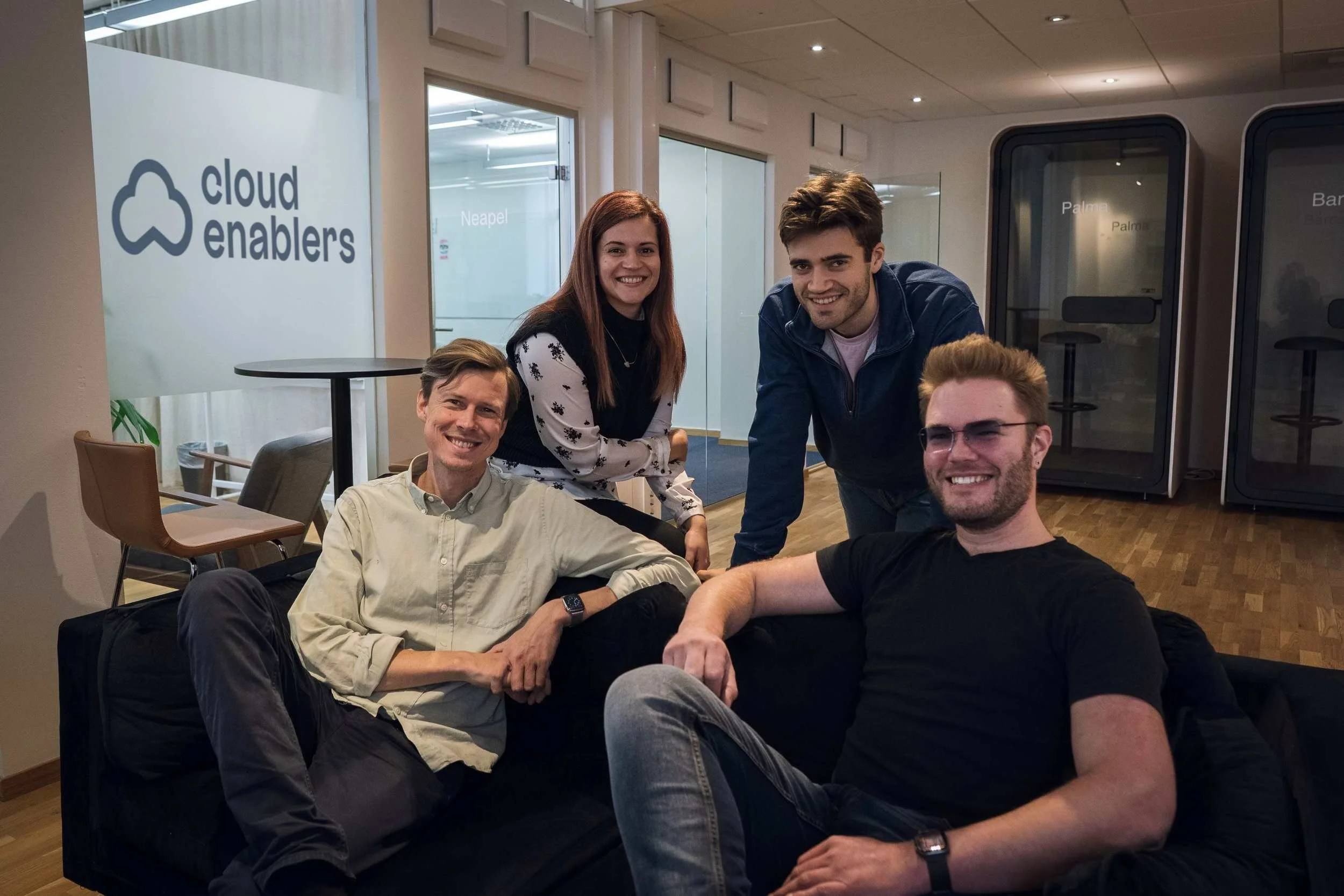 Four young adults smiling in an office lounge, two sitting on a black sofa, two standing behind, with a 'cloud enablers' sign on the wall and office furniture in the background.