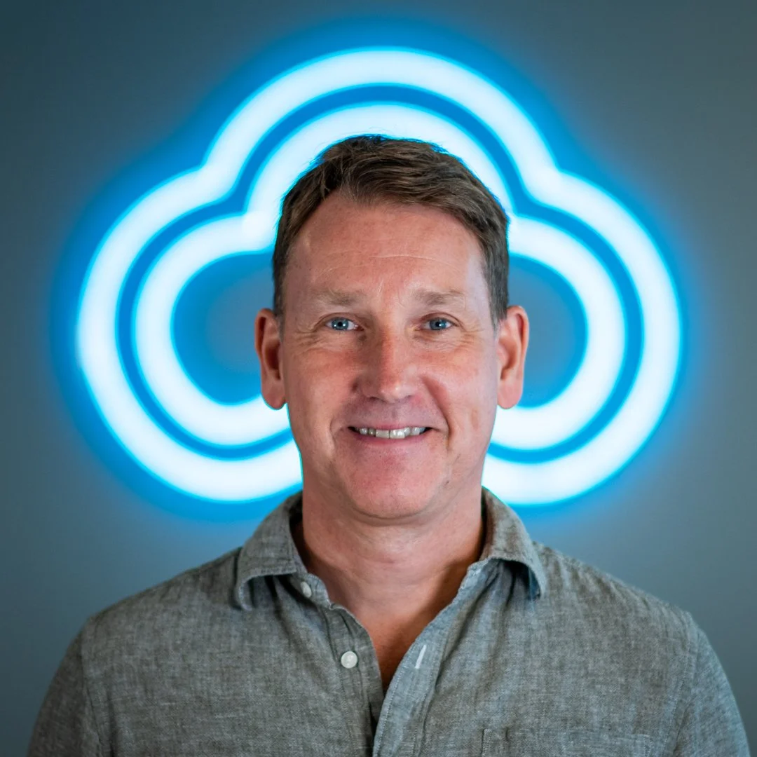 A man with short brown hair and blue eyes smiling, standing in front of a blue cloud-shaped neon sign.