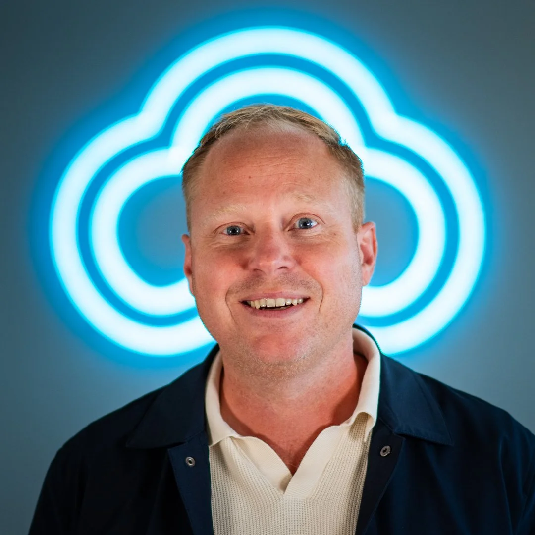 A smiling man with short blond hair, wearing a white collared shirt and dark jacket in front of a blue cloud-shaped neon sign.