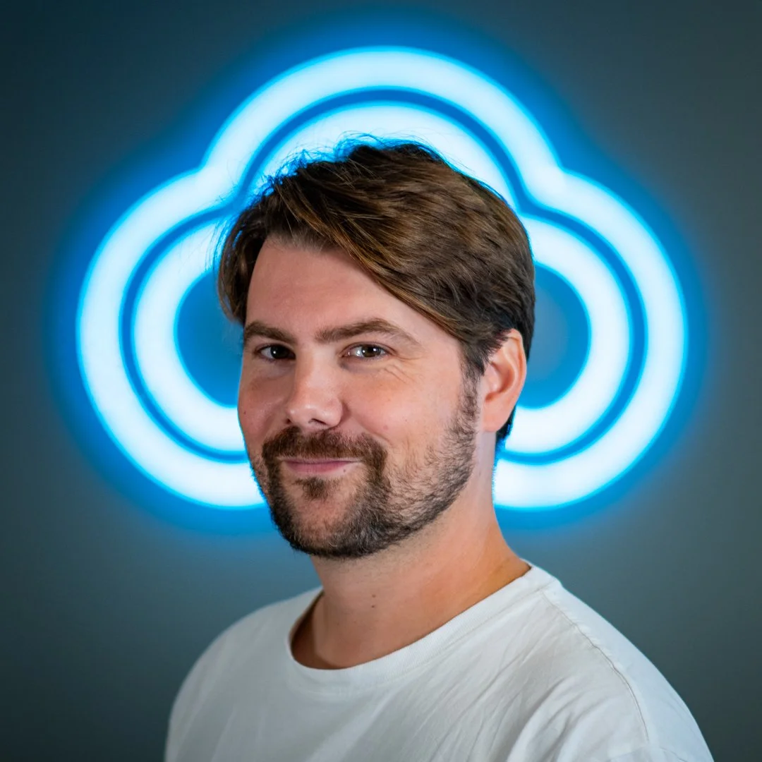 Portrait of a man with brown hair and beard, smiling, standing in front of a blue neon cloud-shaped sign.