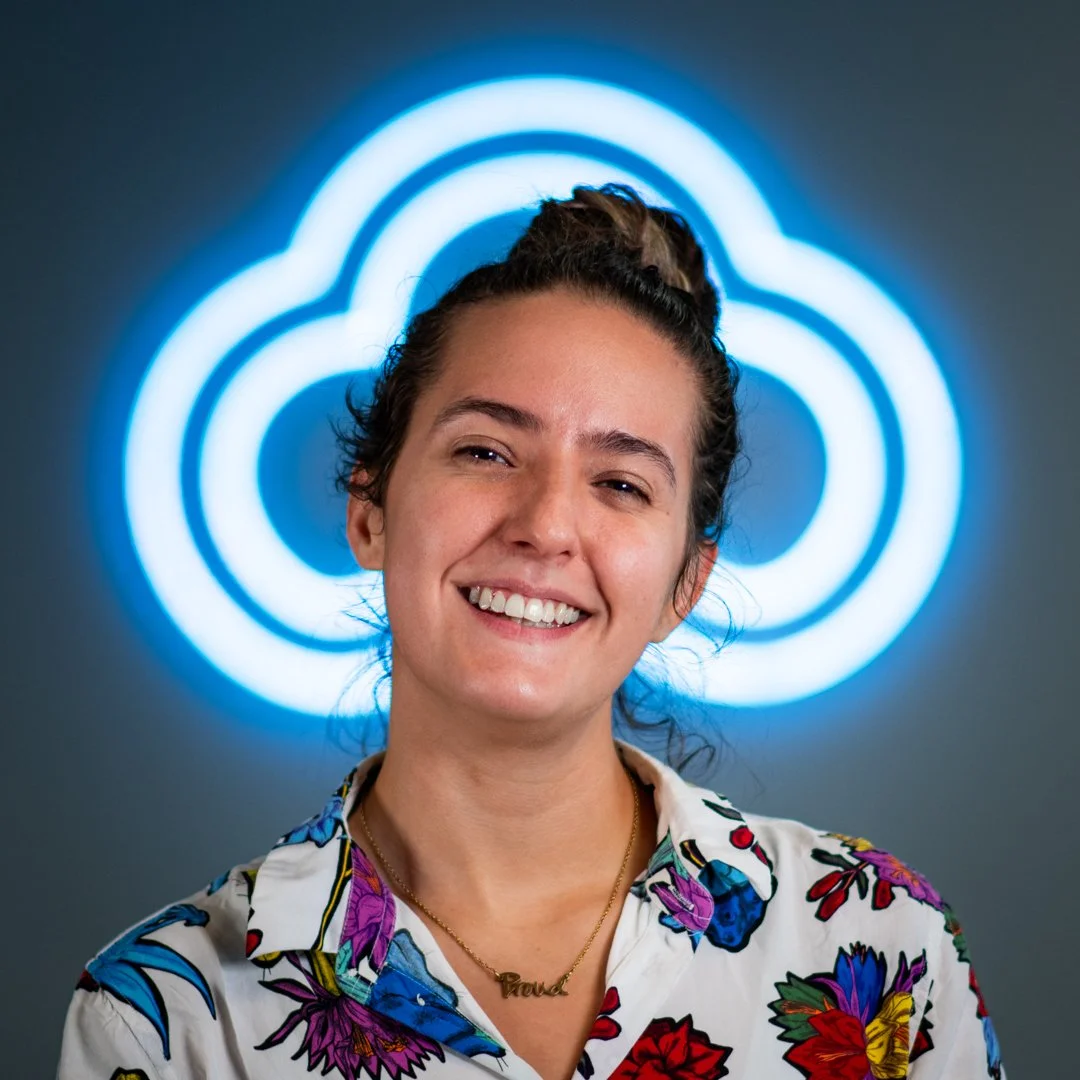 Smiling woman with dark hair tied up, wearing a floral shirt and gold necklace, standing in front of a blue neon cloud logo.