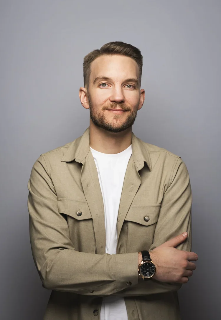 A young man with light brown hair and a beard is smiling slightly, wearing a beige jacket over a white t-shirt, with his arms crossed, standing against a gray background.