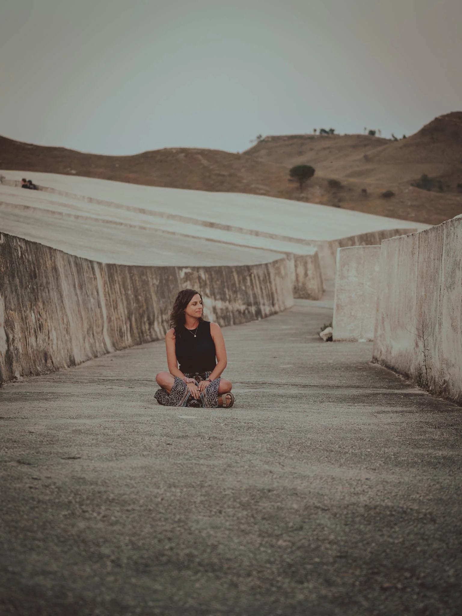 A woman sitting cross-legged on a bridge in a desert landscape with hills in the background.