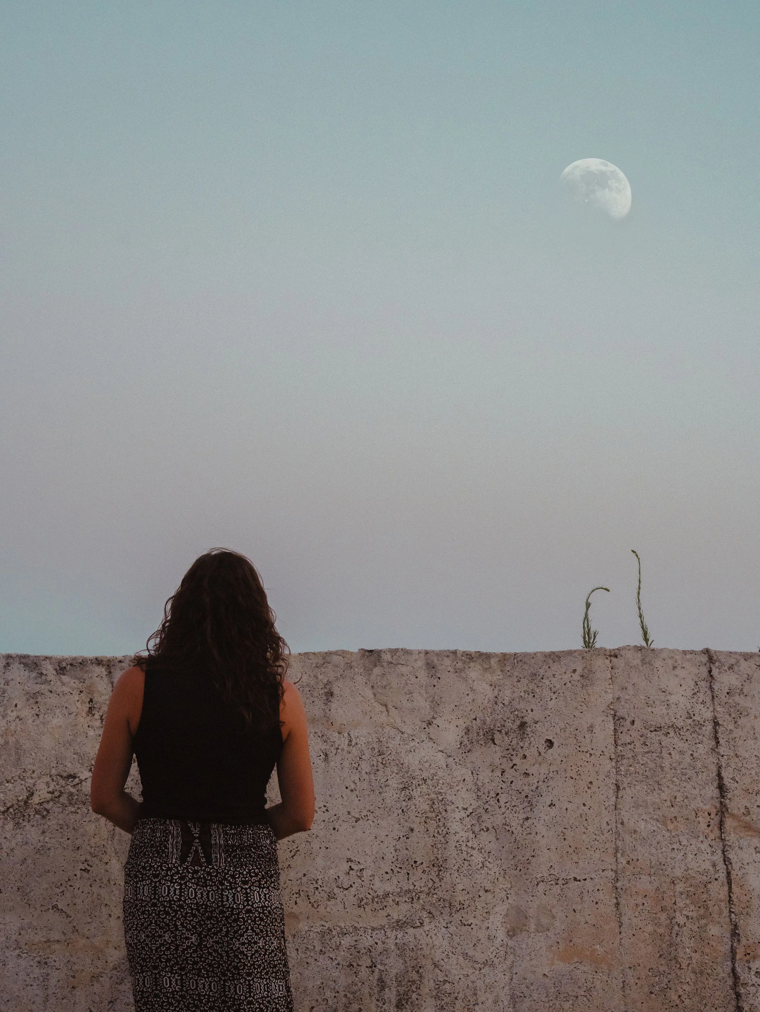 A woman with dark curly hair stands facing a concrete wall, looking at the rising moon in the early evening sky.