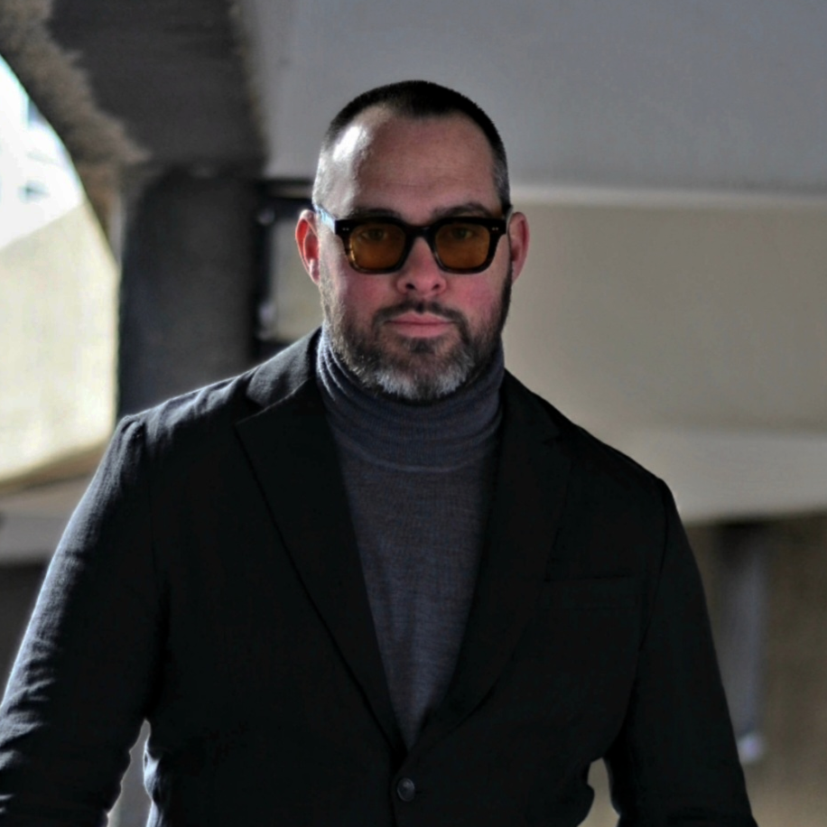 A man with a beard wearing glasses, a dark blazer, and a turtleneck, standing indoors.