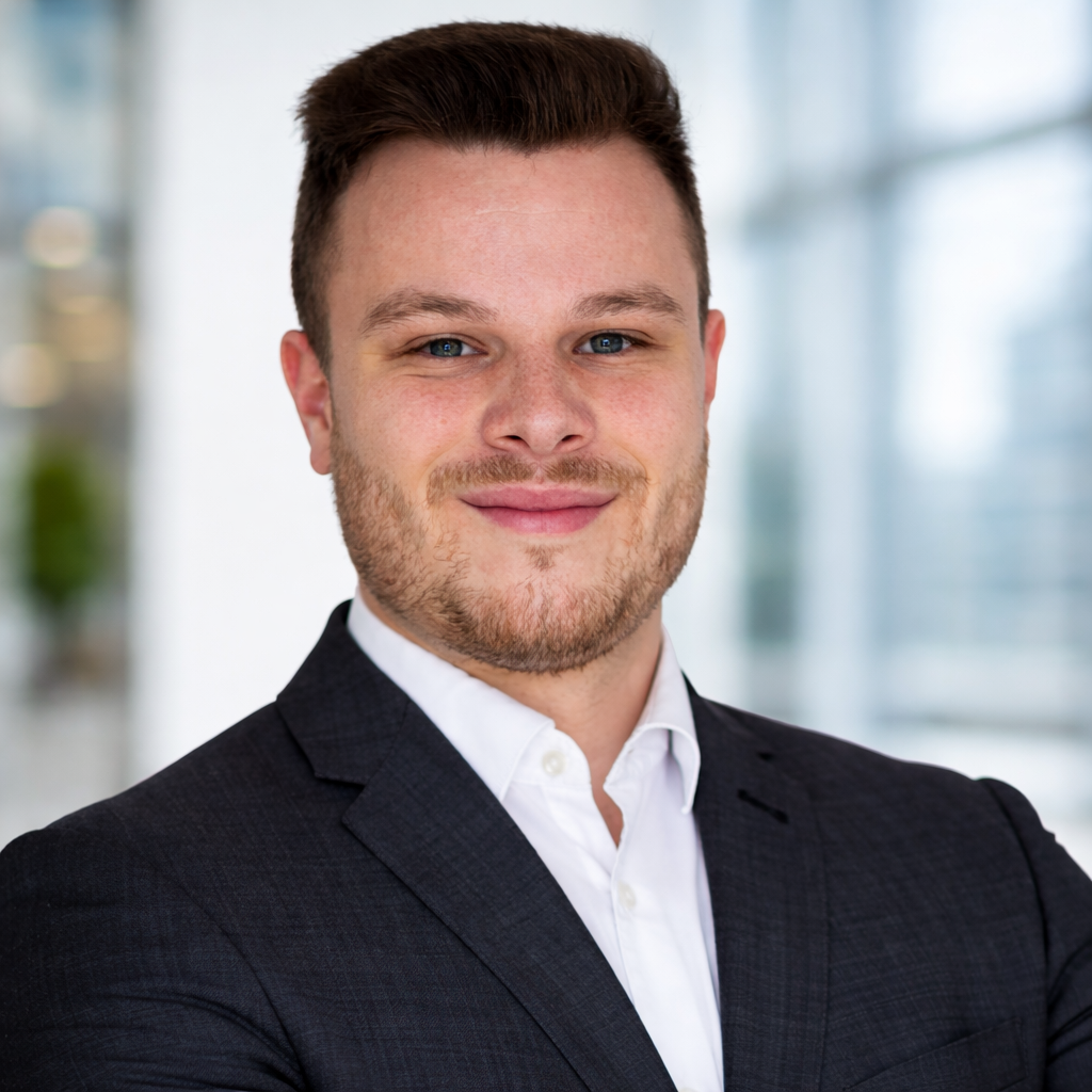 Ein Mann in einem schwarzen Anzug und weißen Hemd, lächelnd, vor einem modernen Bürogebäude