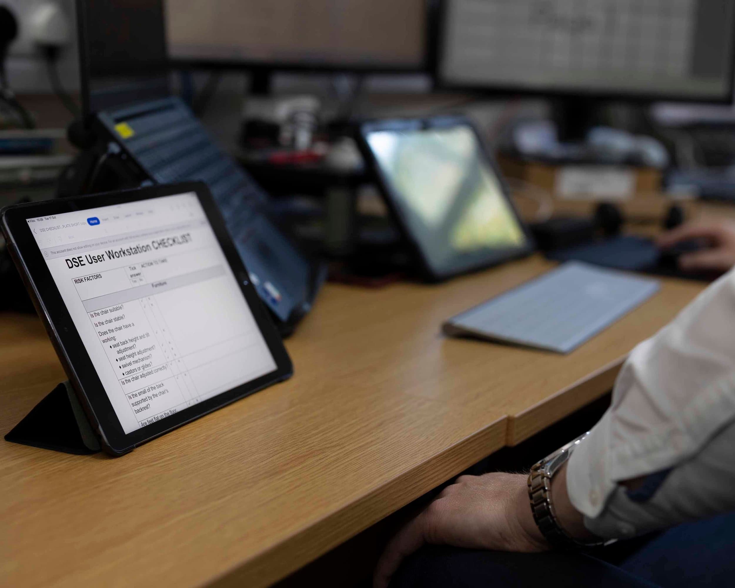 A person sitting at a wooden desk with digital devices including a tablet displaying a checklist, multiple computer monitors, and smartphones. The person is wearing a white shirt and a wristwatch.