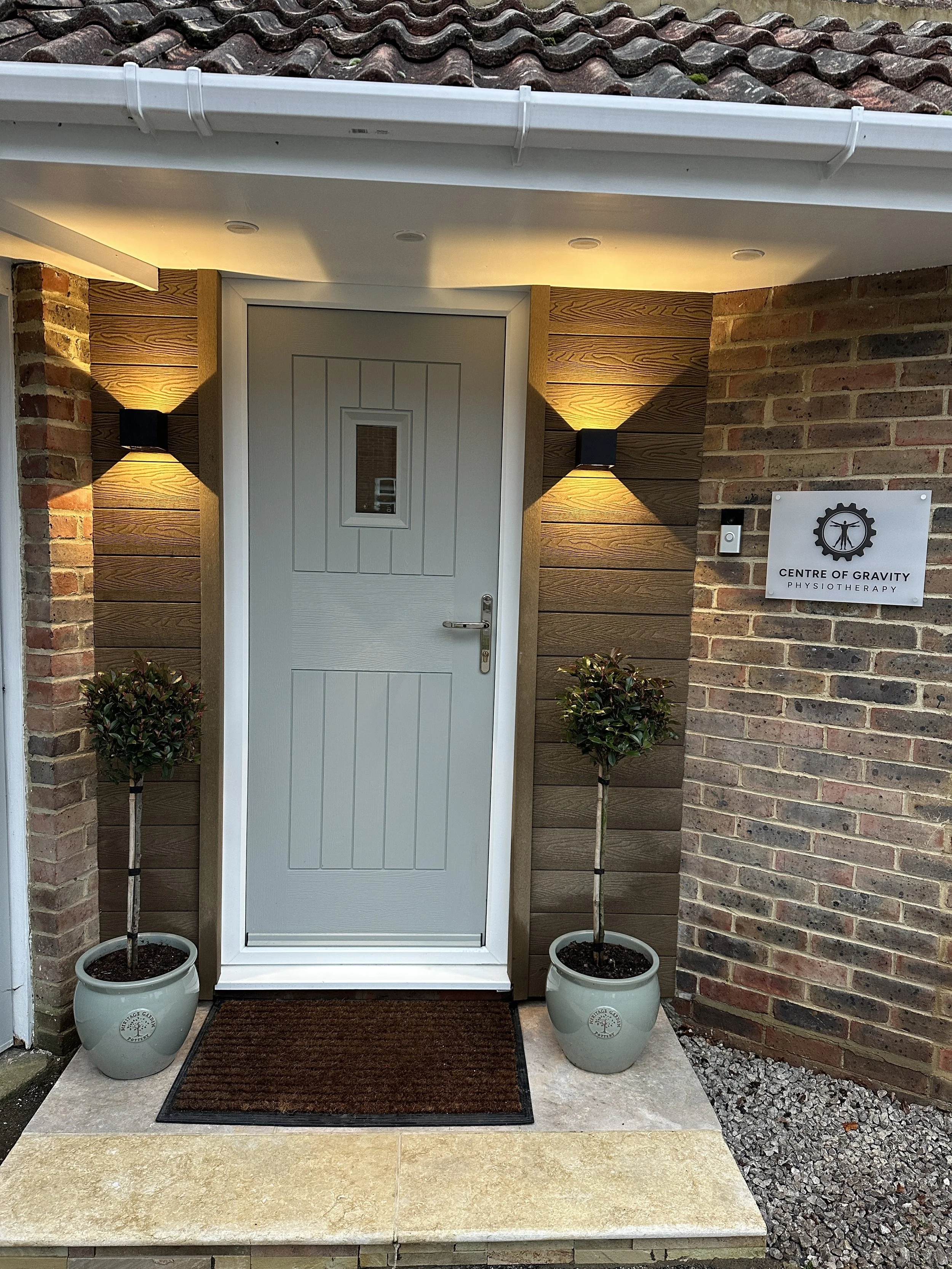 The entrance of a physiotherapy clinic named 'Centre of Gravity Physotherapy' with a gray door, two potted trees on each side, a doormat, wall-mounted lights, and a sign on the brick wall.