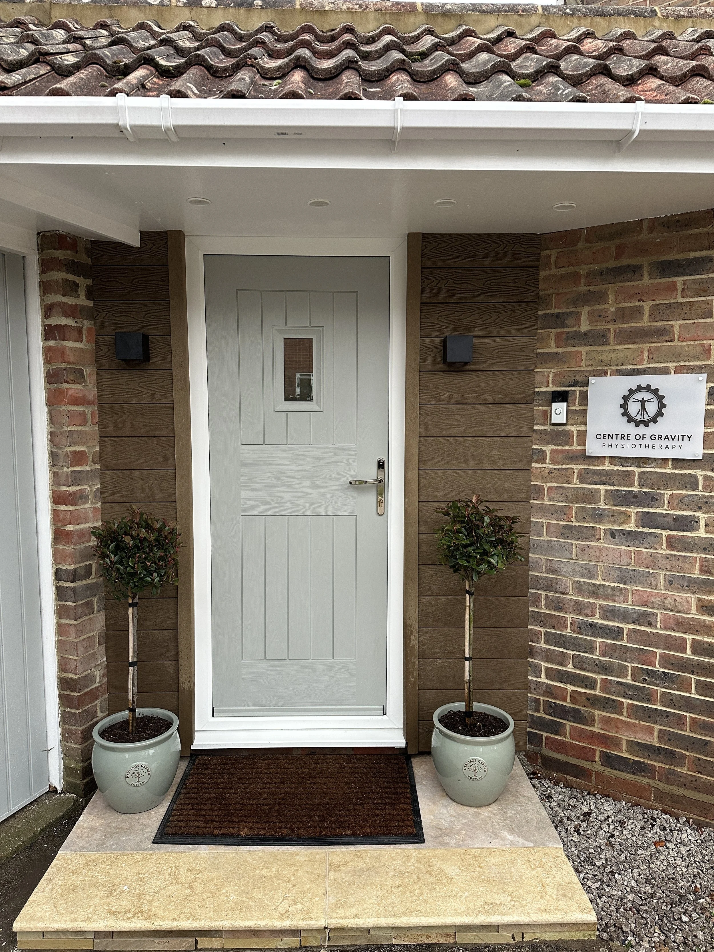 Front entrance of a building with a white door, two potted trees on either side, a brown doormat, a brick wall to the right with a sign for Centre of Gravity Physiotherapy, and an overhanging roof with tiles.