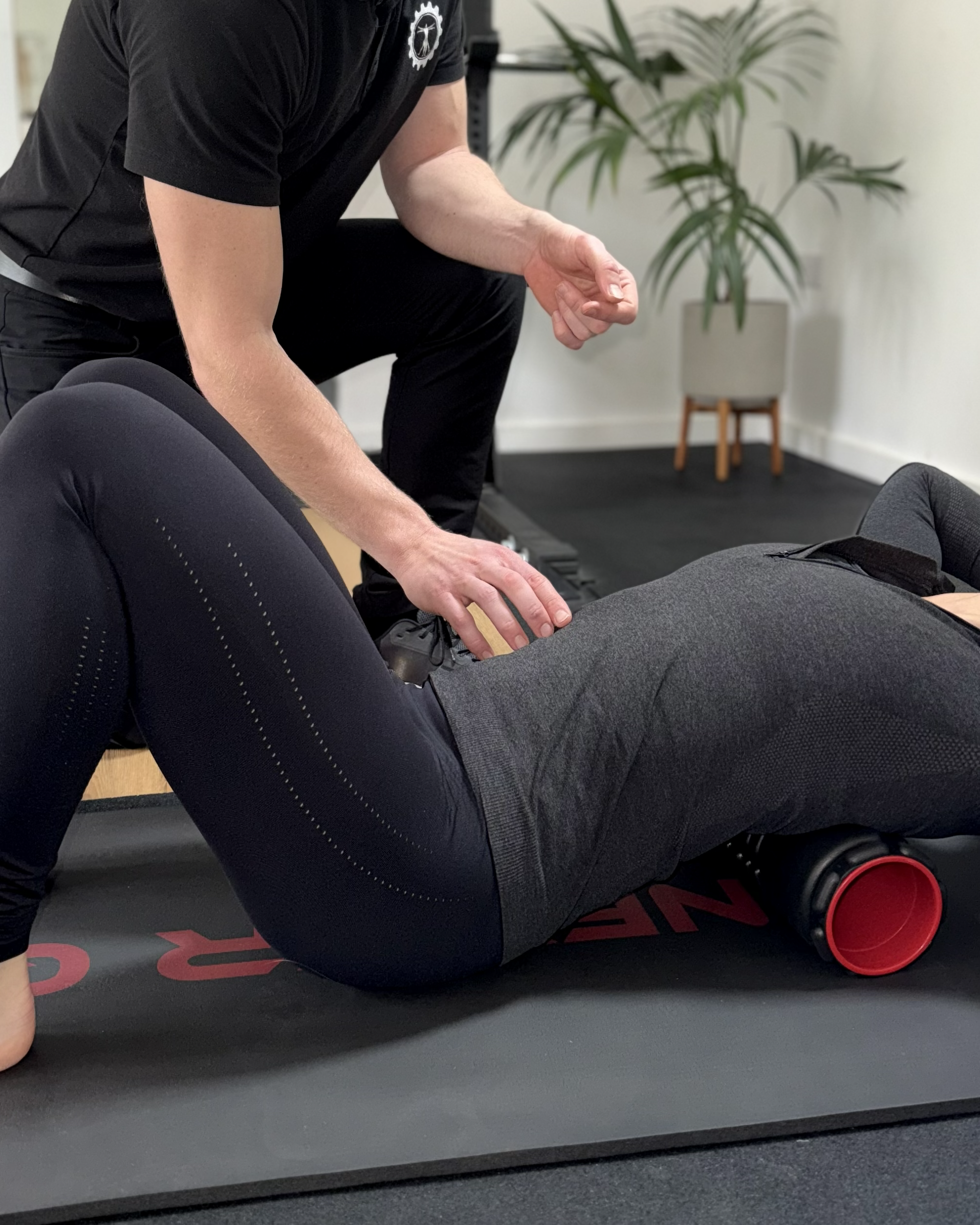 A person receiving physical therapy or massage on their lower back while lying face down on a black exercise mat. The person providing therapy is wearing a black shirt and black pants, and is kneeling on the mat. There is a green potted plant in the background and a piece of gym equipment in the corner.