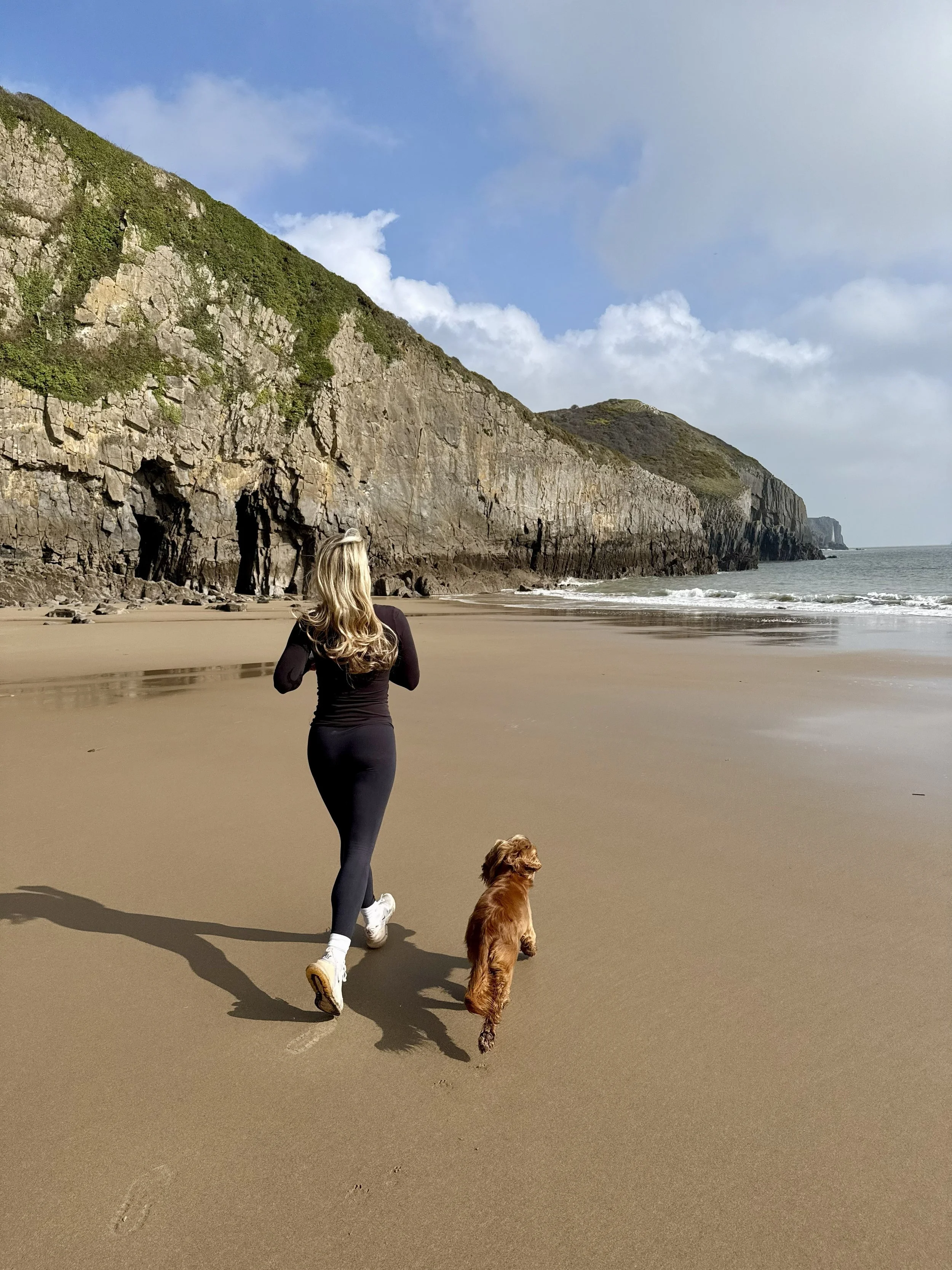 A woman running on a sandy beach with a golden retriever dog. The background features tall green cliffs and a partly cloudy sky.