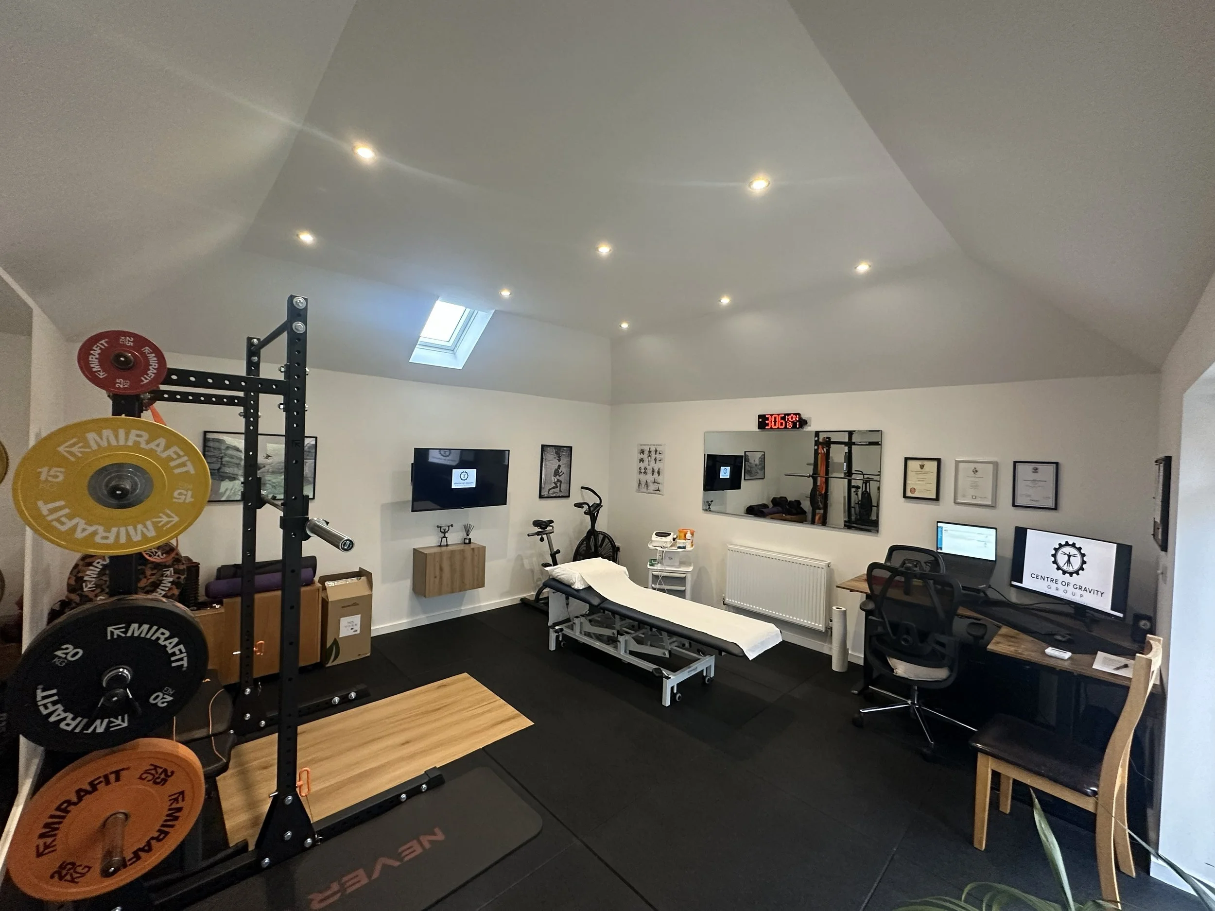 A fitness and therapy room with weightlifting equipment, a massage table, exercise bike, computers, and framed certificates on the wall.