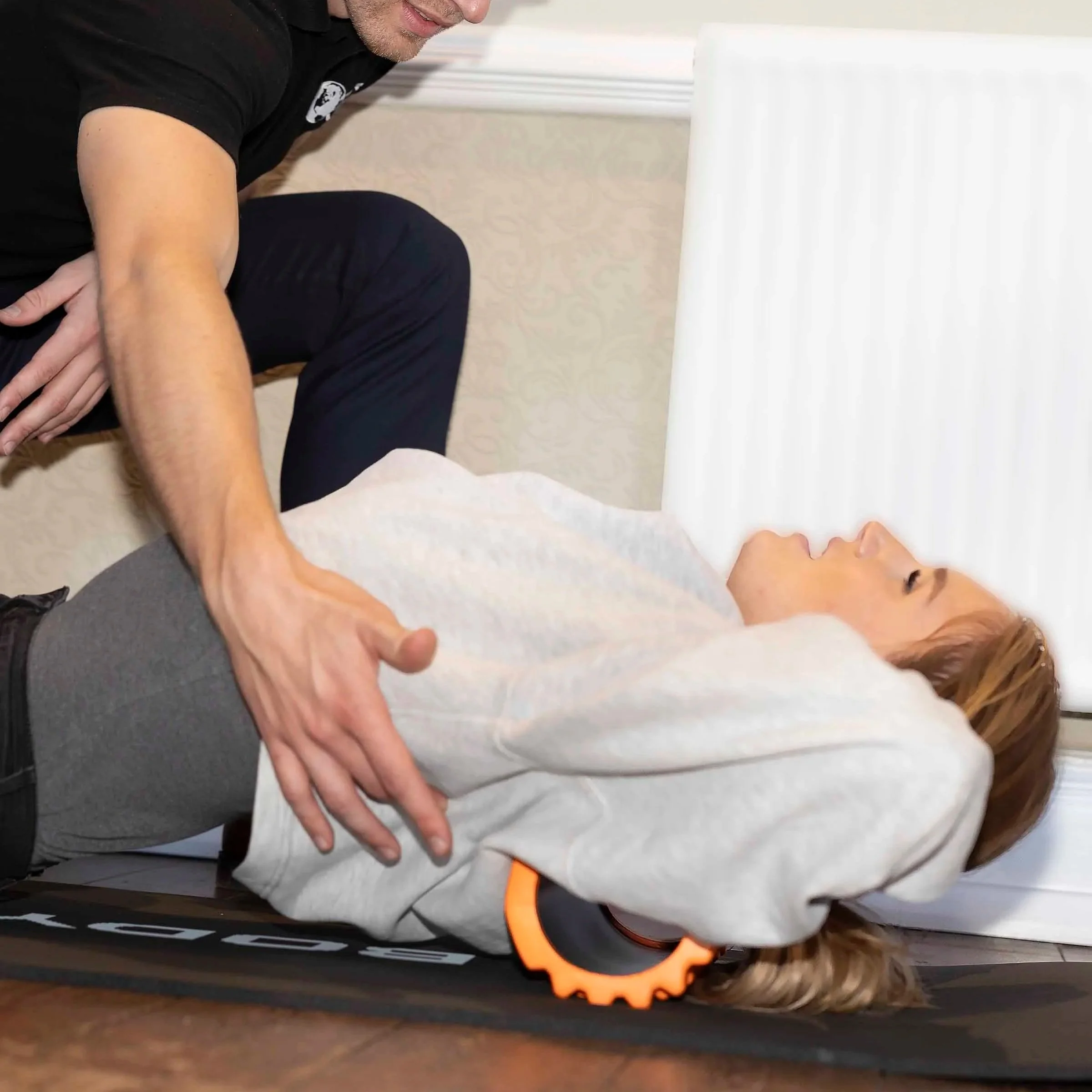 A person practicing a rescue drill on a woman lying on the ground with a foam roller under her head. The woman is wearing a gray hoodie, and the man is wearing a black shirt, kneeling near her.