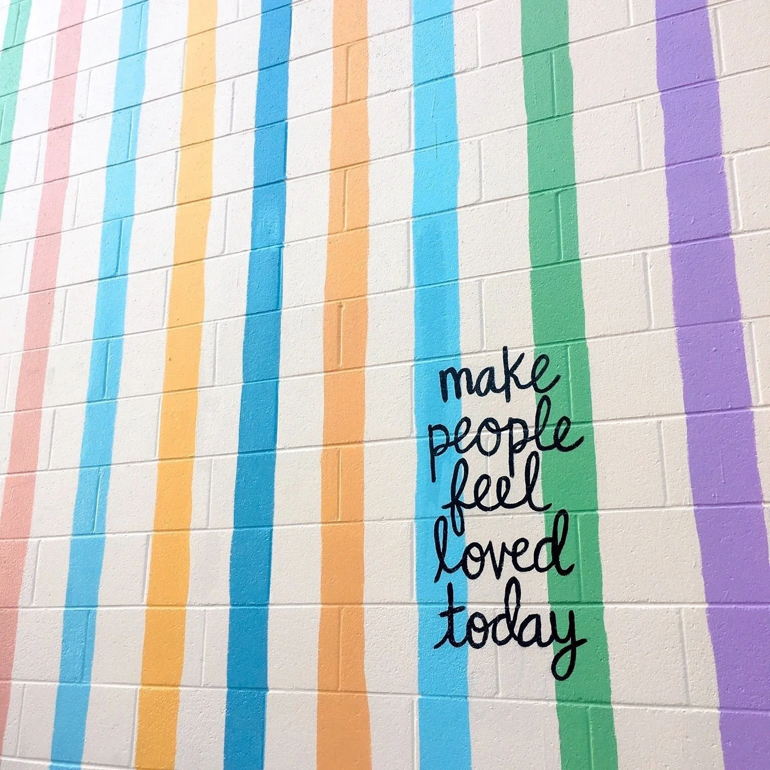 A colorful mural with vertical pastel stripes in pink, yellow, blue, green, and purple. Handwritten text in black reads: 'make people feel loved today'.
