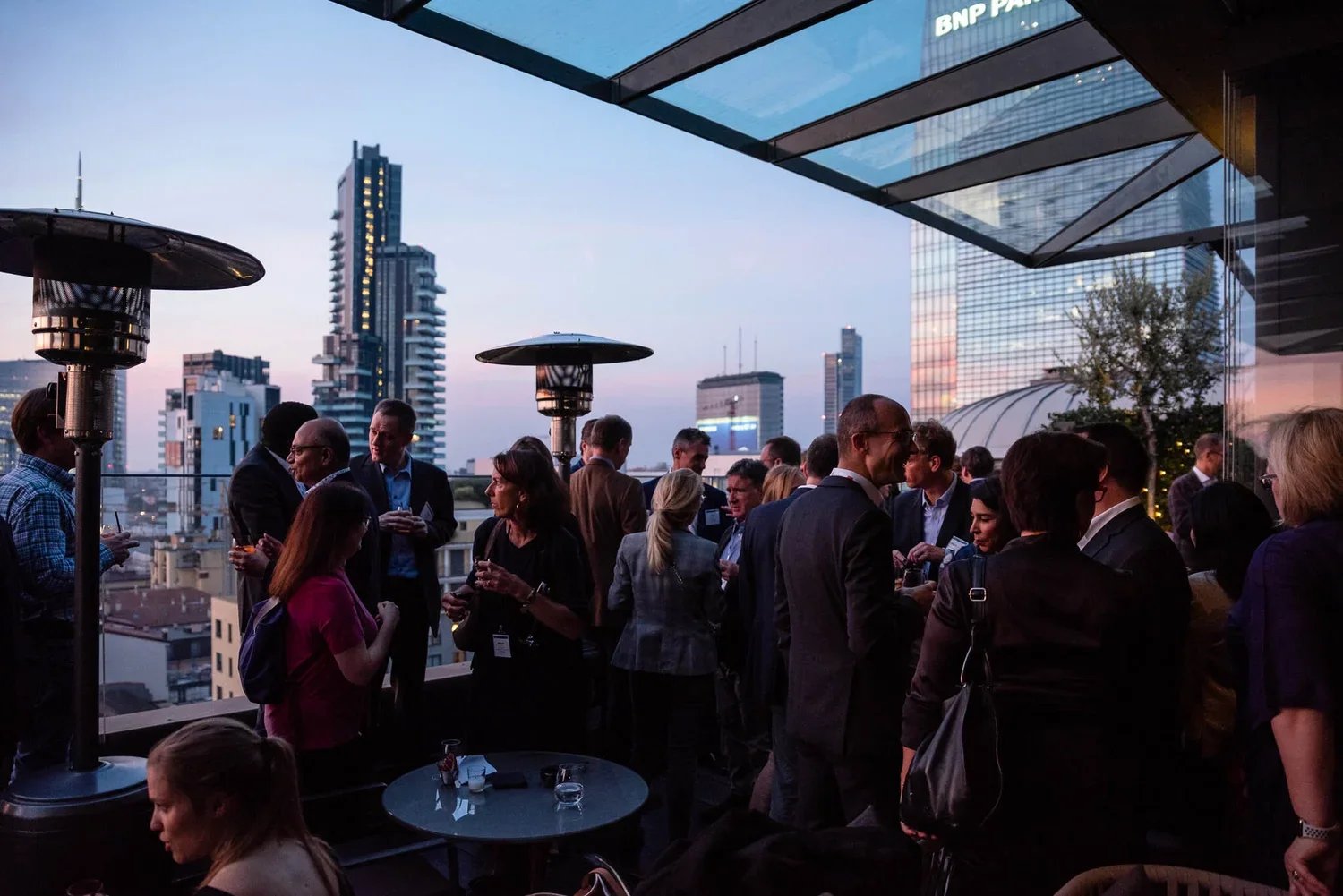 People socializing on a rooftop terrace in a city at dusk with modern skyscrapers in the background.