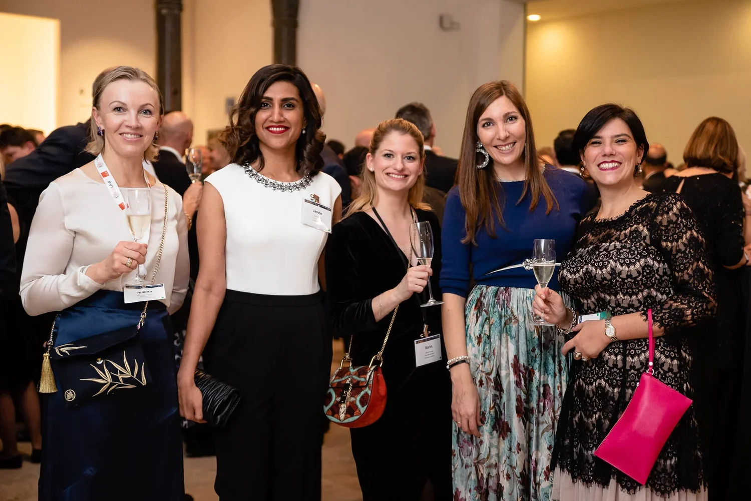 Five women smiling at a social event, holding champagne glasses. They are dressed in formal attire, with name tags visible, and are standing close together in a crowded indoor venue.