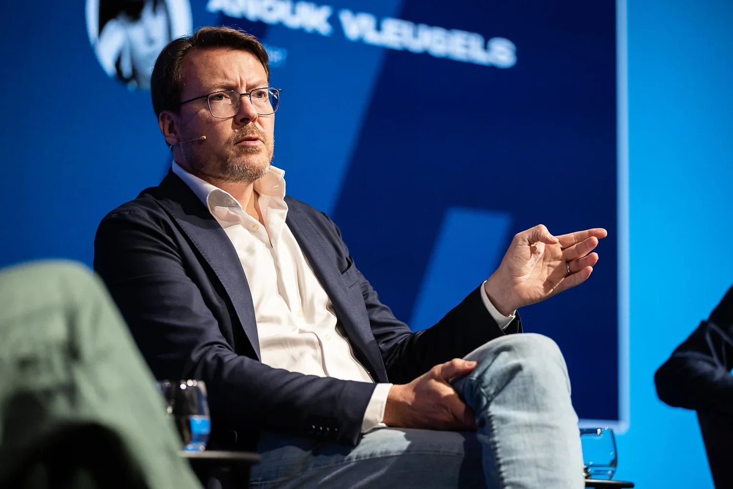 A man with glasses, a white shirt, dark blazer, and jeans is speaking during a panel discussion. He has a beard and is gesturing with his right hand while seated on a stage.