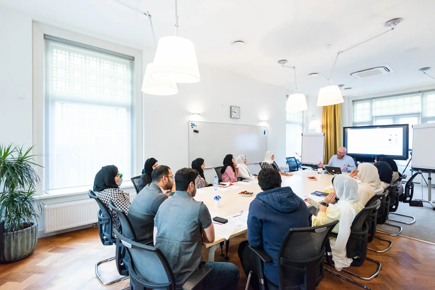 A diverse group of professionals attending a meeting in a bright conference room with large windows, a whiteboard, and a large screen at the front, listening to a presenter.