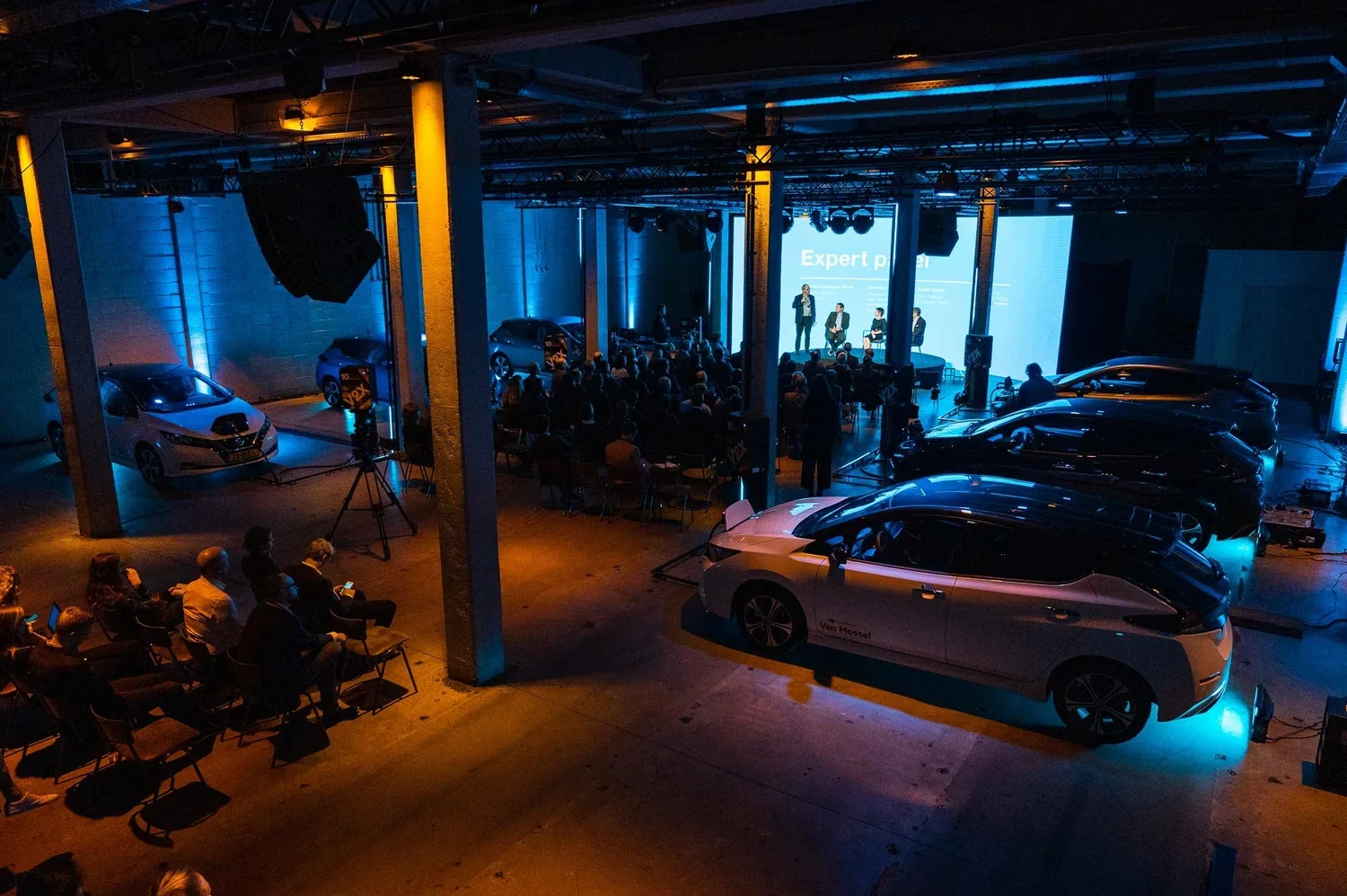 An indoor car showroom or event space with multiple electric cars displayed, a stage with a panel discussion or presentation, and audience seated and engaging with the event, illuminated with blue and orange lighting.