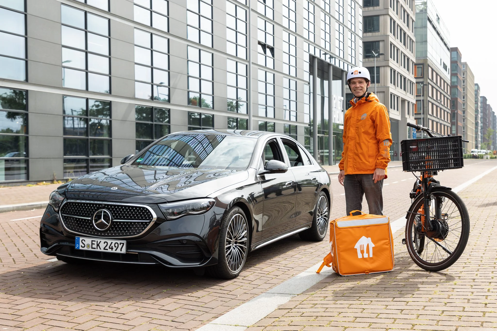 A delivery person standing next to a Mercedes-Benz car with a bicycle and a delivery bag on the sidewalk in an urban area.