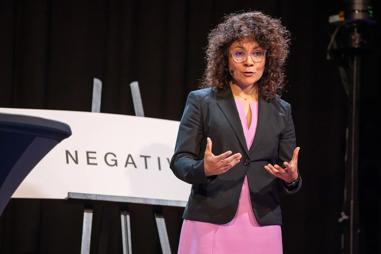 woman with curly hair speaking on stage in front of a sign that reads 'NEGATIVE' in black letters, wearing glasses, a black blazer, and a pink dress.