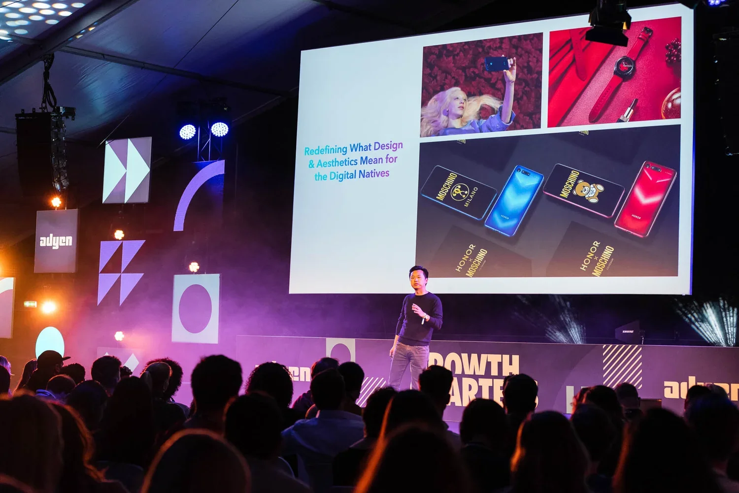 A speaker at a conference discussing digital design and aesthetics, with a large screen behind him displaying images of smartphones and accessories, and an audience watching in a dimly lit room with colorful stage lighting.