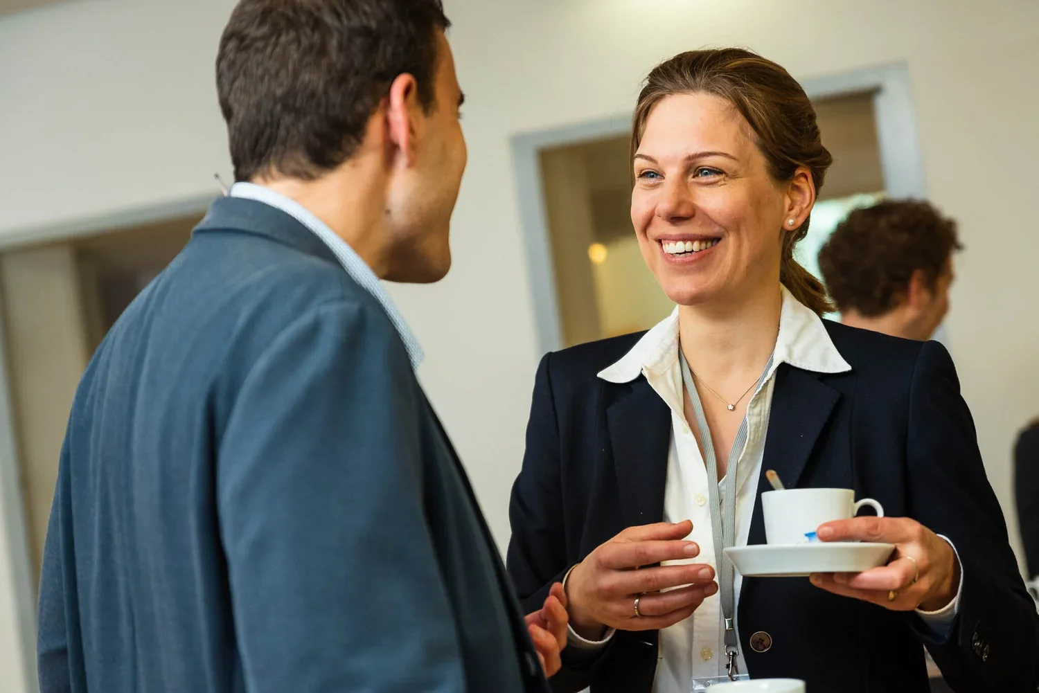 A woman with brown hair smiling and holding a coffee cup and saucer, engaging in conversation with a man in a suit at a professional event.