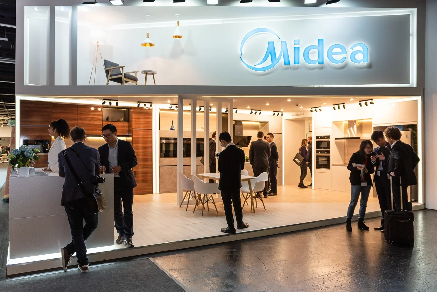 An exhibition booth for Midea, showcasing kitchen appliances and home furnishings, with several visitors and representatives engaging in conversations.