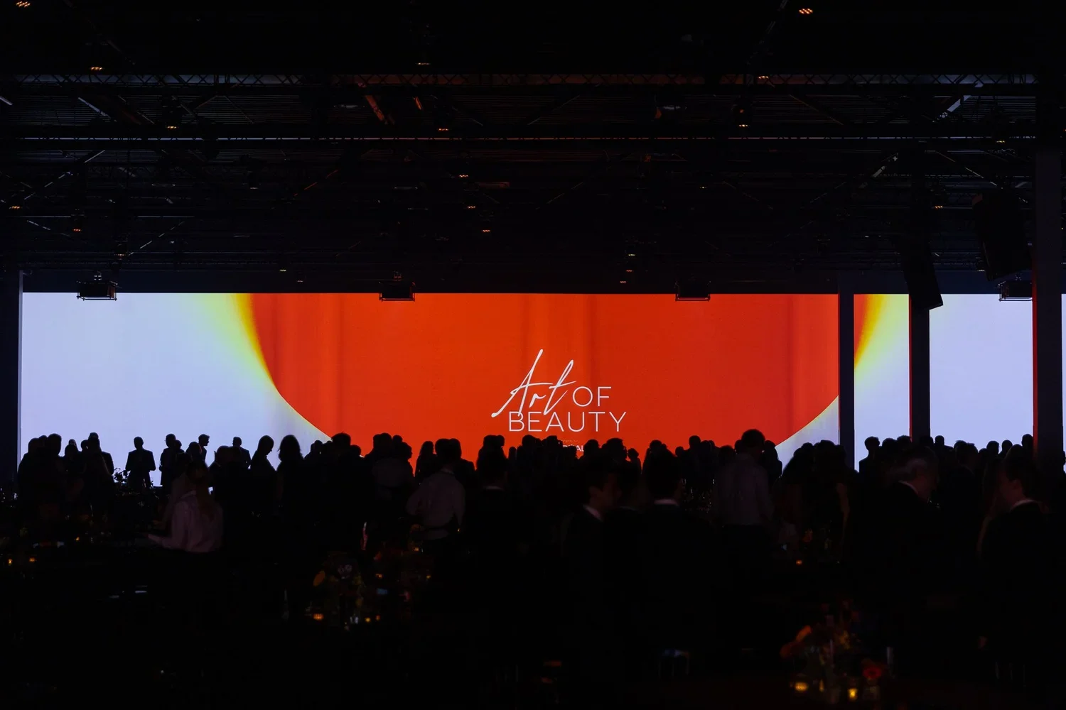 A large indoor event with a darkened room and a big screen displaying the words 'Art of Beauty' on a red and white abstract background. There are many people seated and standing, facing the screen.