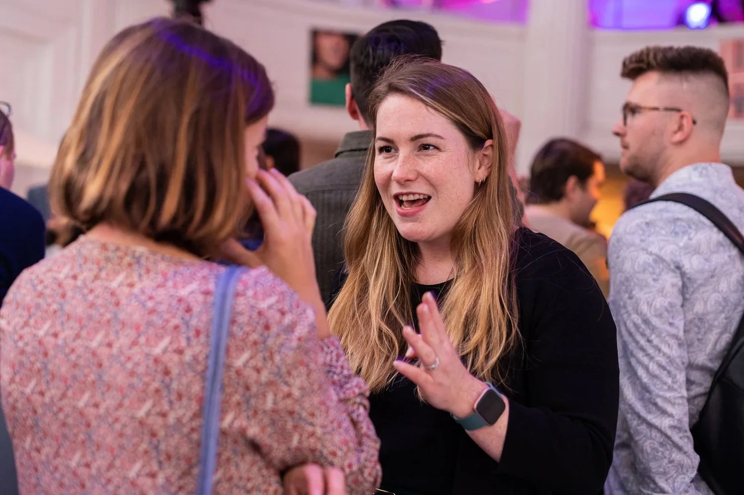 Two women are engaged in a conversation at a social event. One woman, with light brown hair, wearing a dark top, is smiling and gesturing with her hand. The other woman, with reddish-brown hair, wearing a patterned top, is listening. The background s