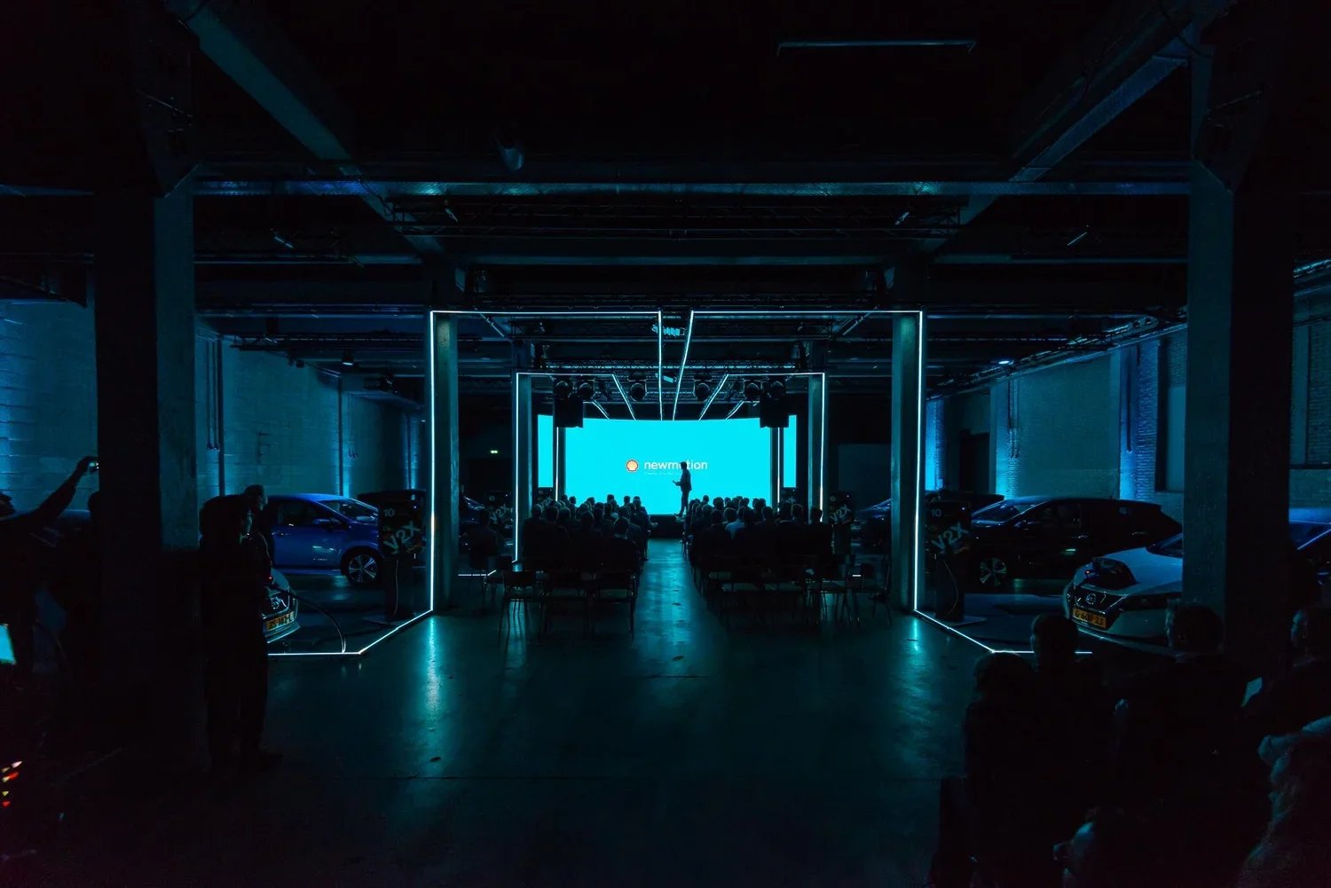A dark indoor event space with a presentation stage featuring a large blue screen that displays the logo and name "newmotion." The room has a modern industrial design with exposed ceiling pipes and walls. There are cars parked along the sides and peo