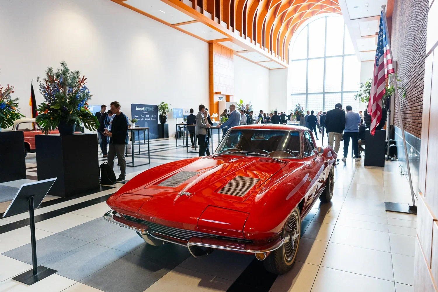 A red vintage sports car on display at an indoor automobile exhibition with people in the background. The setting features a large glass window and American flag on the right side.